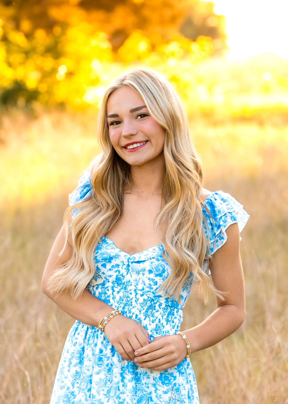 Young woman in blue floral dress smiling in golden field with warm autumn sunlight background.