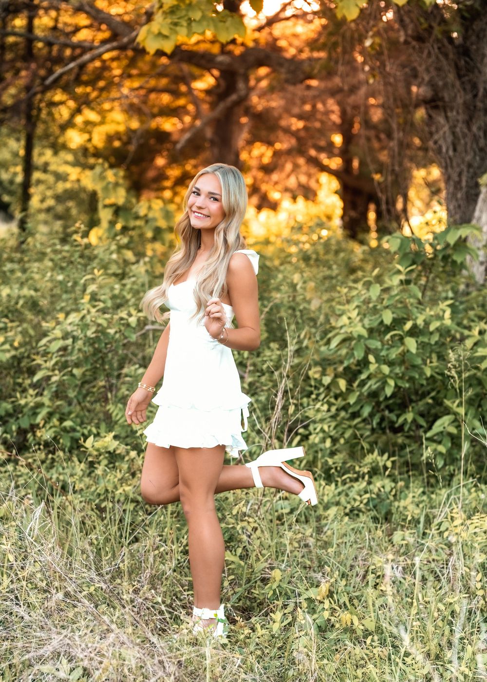 Young woman in white mini dress posing playfully in lush green field at sunset.
