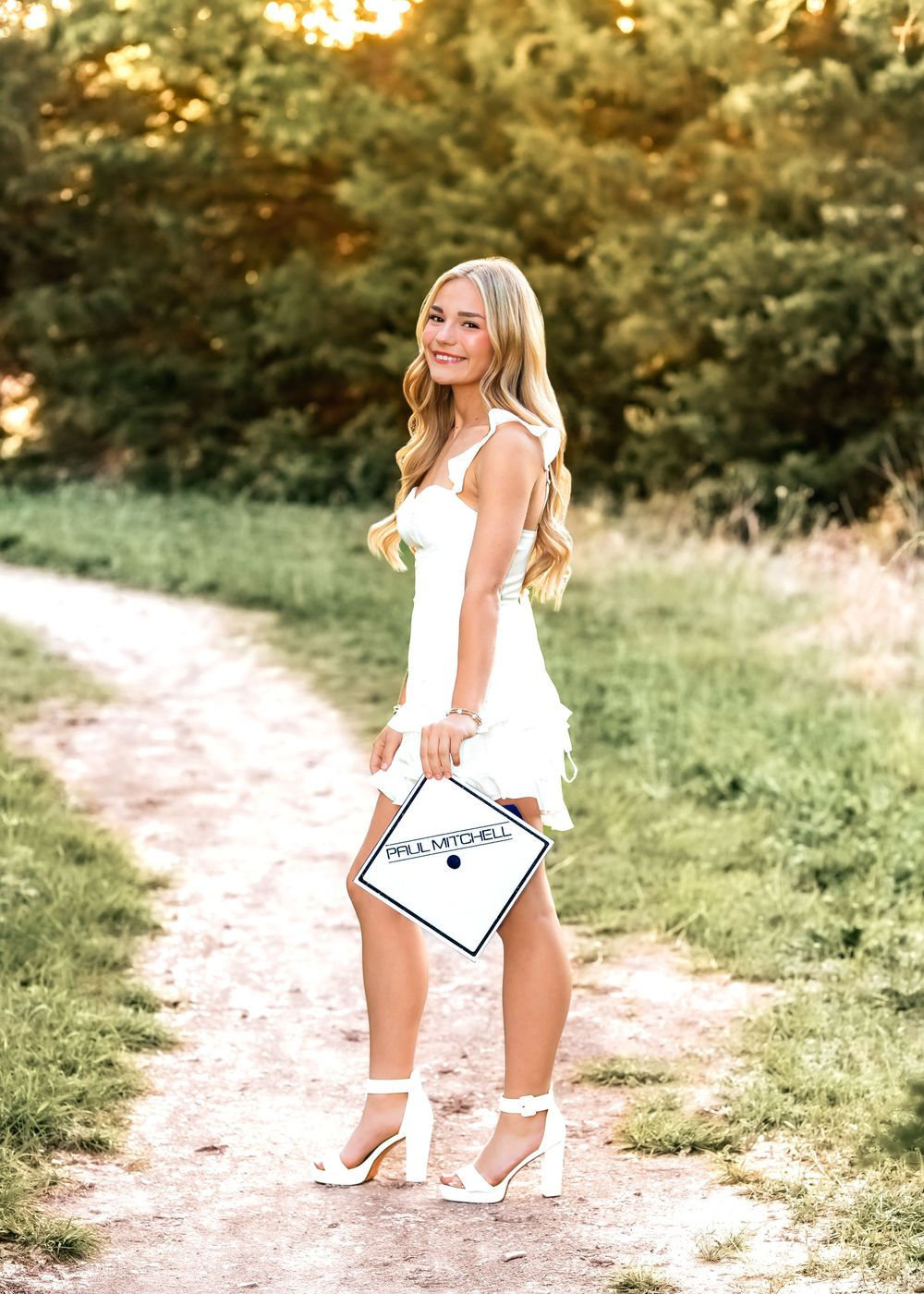 Young woman in white dress holding graduation cap on a scenic dirt path surrounded by lush green trees.
