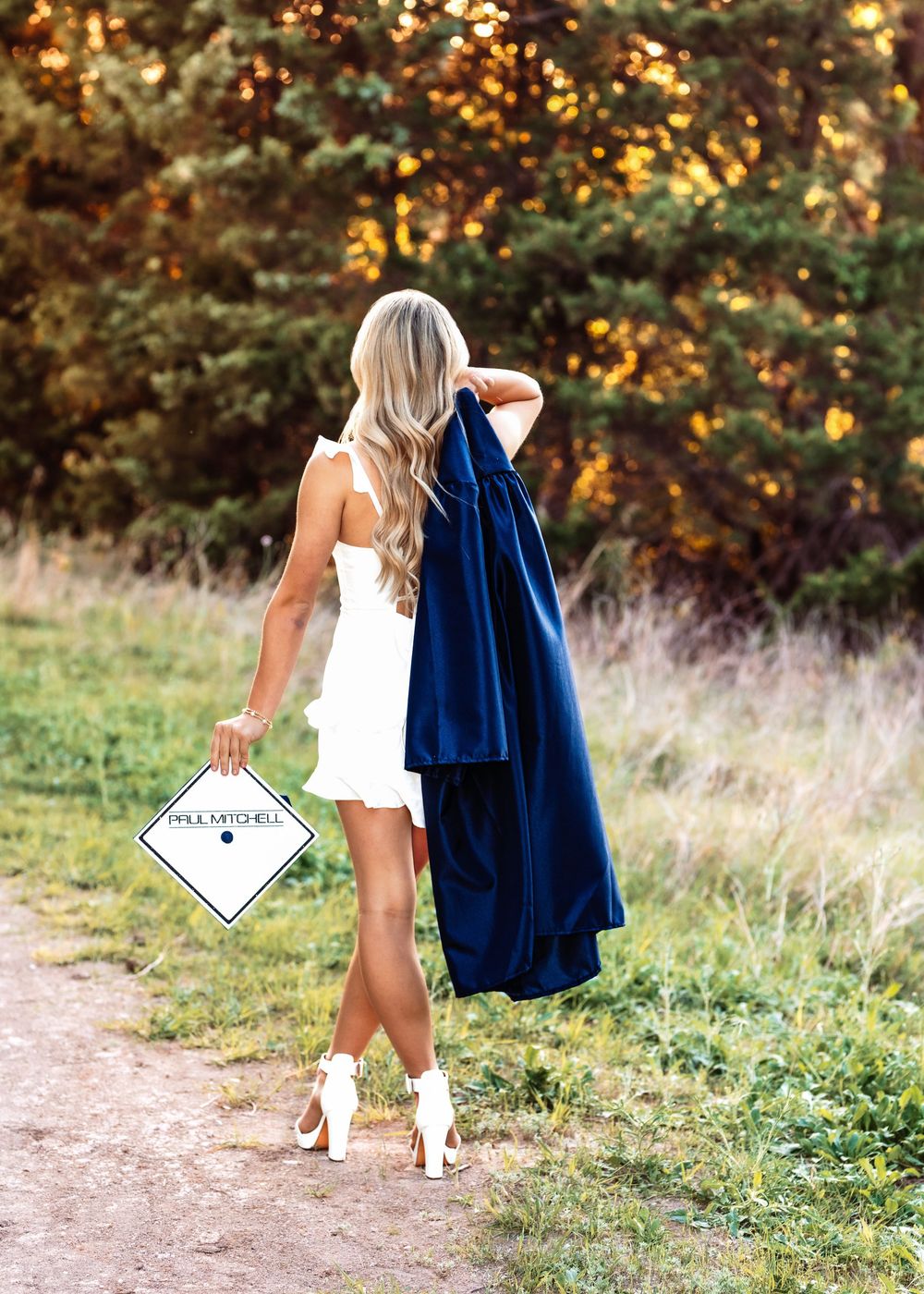Blonde woman in white dress holding navy graduation gown and cap outdoors on a dirt path surrounded by trees.