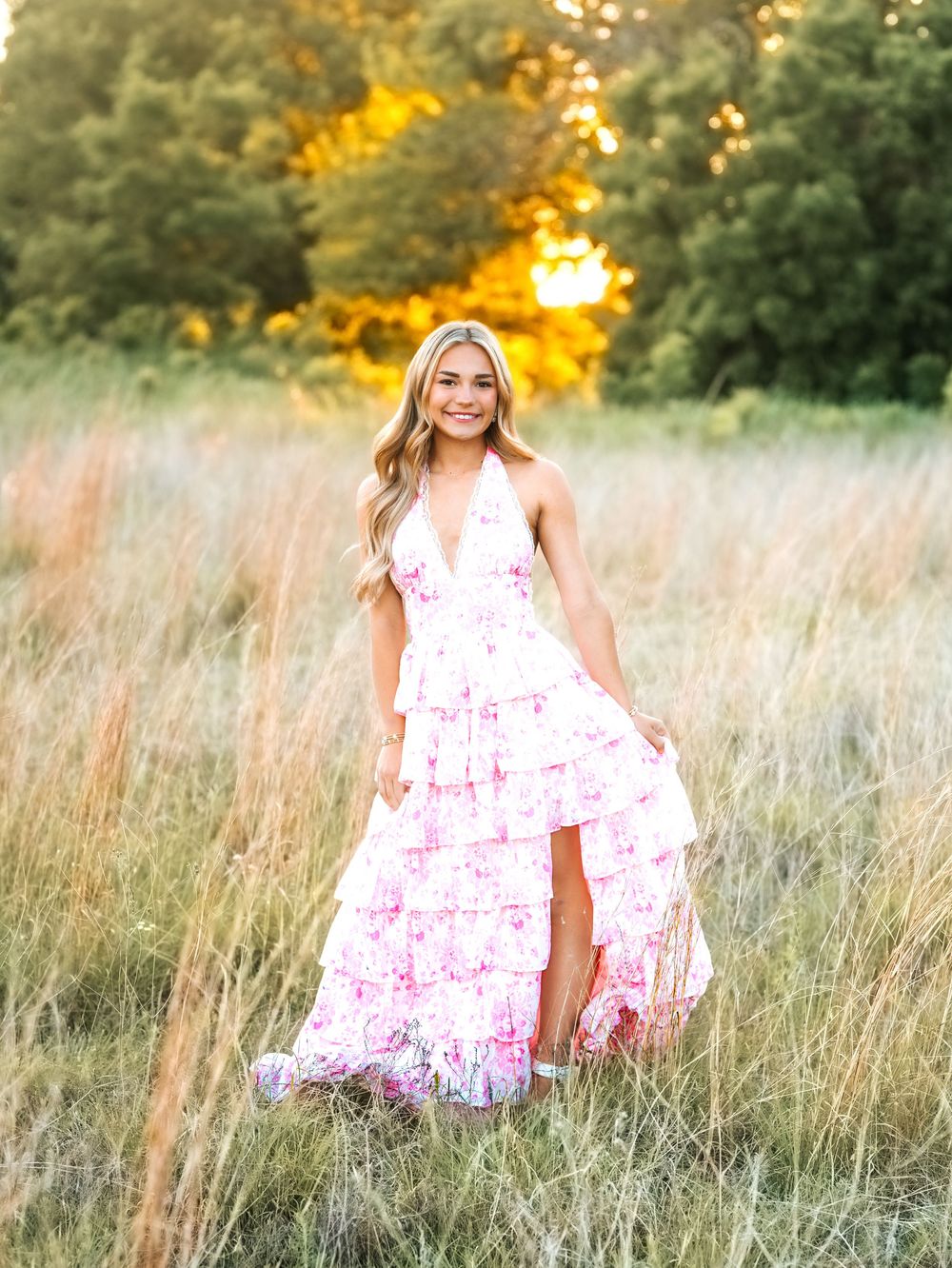 Blonde woman in pink floral halter maxi dress standing in golden field at sunset.