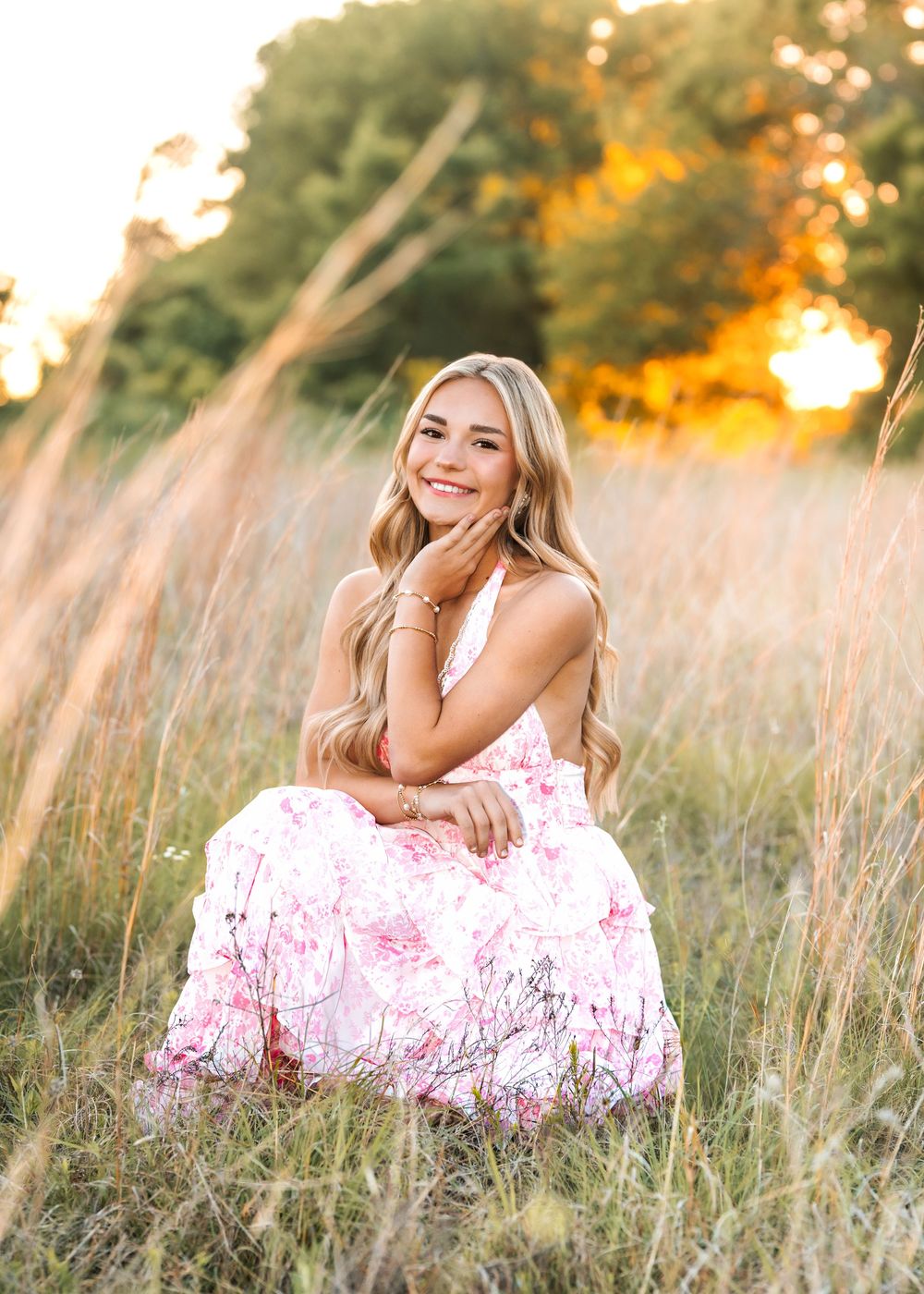 Young woman in floral pink dress sitting in golden field at sunset, smiling warmly with long blonde hair.