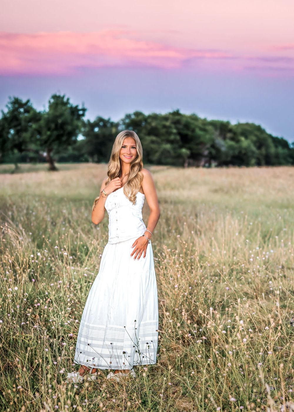 Blonde woman in white maxi dress standing in golden field at sunset with pink sky and green trees.