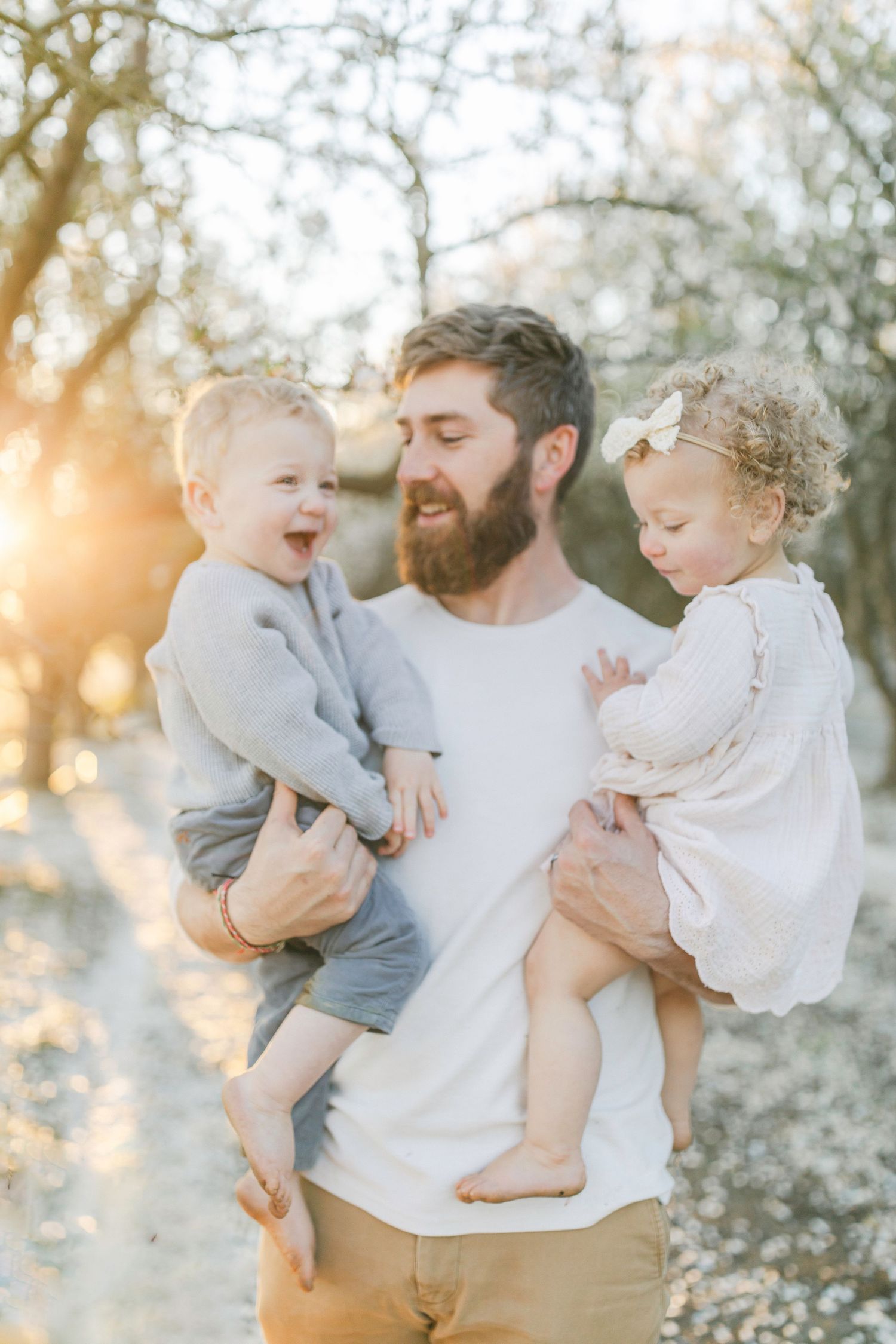Bearded father holding two toddlers outdoors at golden hour, smiling joyfully among blooming trees in soft natural light.