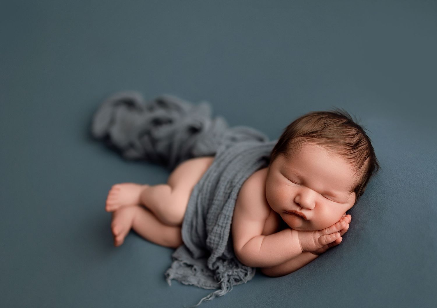 Newborn baby sleeping peacefully wrapped in a gray swaddle on a teal blue backdrop in a professional photo.