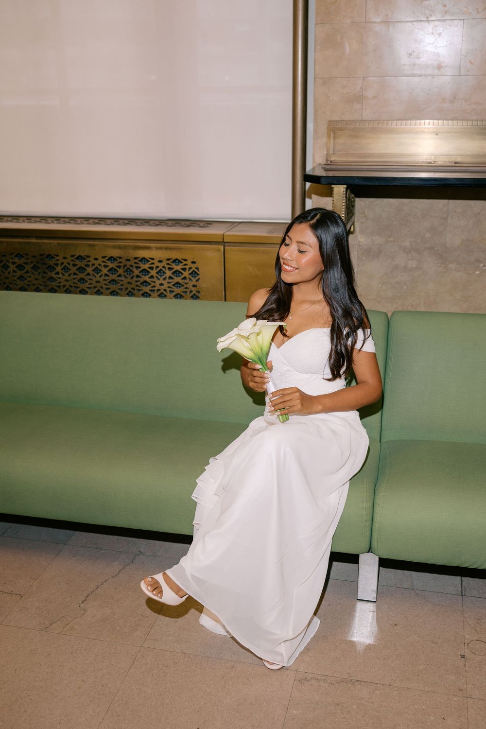 Bride in white off-shoulder gown sits on green bench holding calla lily bouquet, smiling softly.