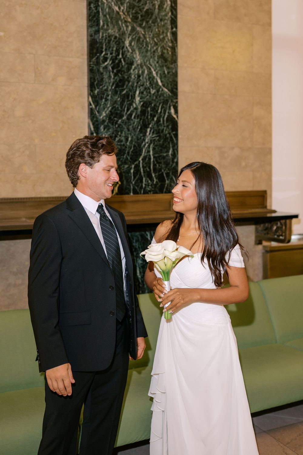 Bride in white dress holding calla lilies smiles at groom in black suit during elegant indoor wedding ceremony.