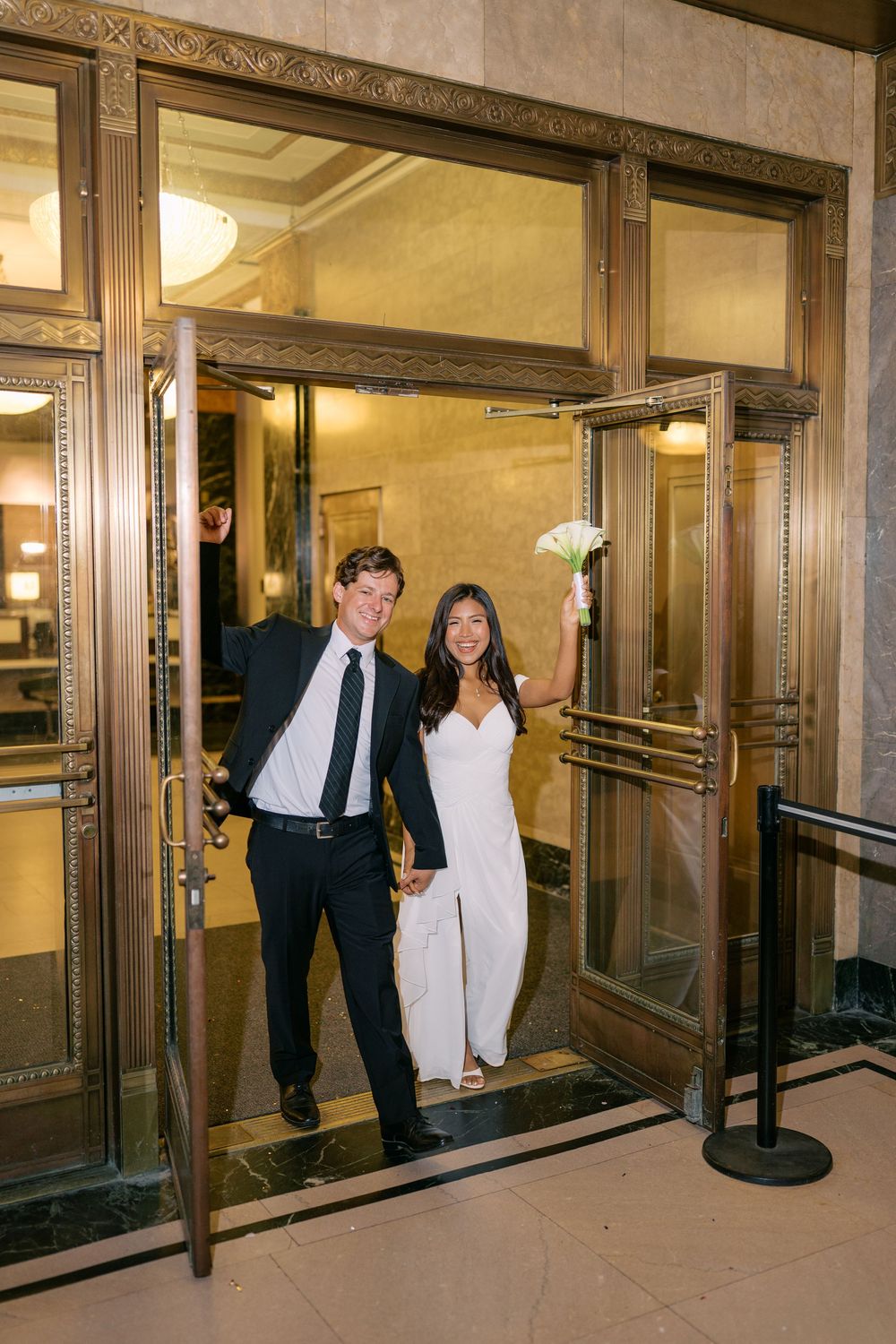 Joyful couple in wedding attire celebrating at ornate golden doors, bride in white jumpsuit holding flowers.