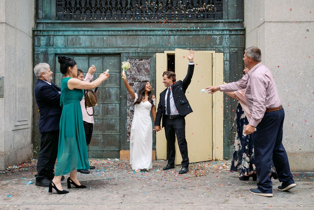 Wedding couple celebrates outside with guests throwing confetti near ornate bronze doors.