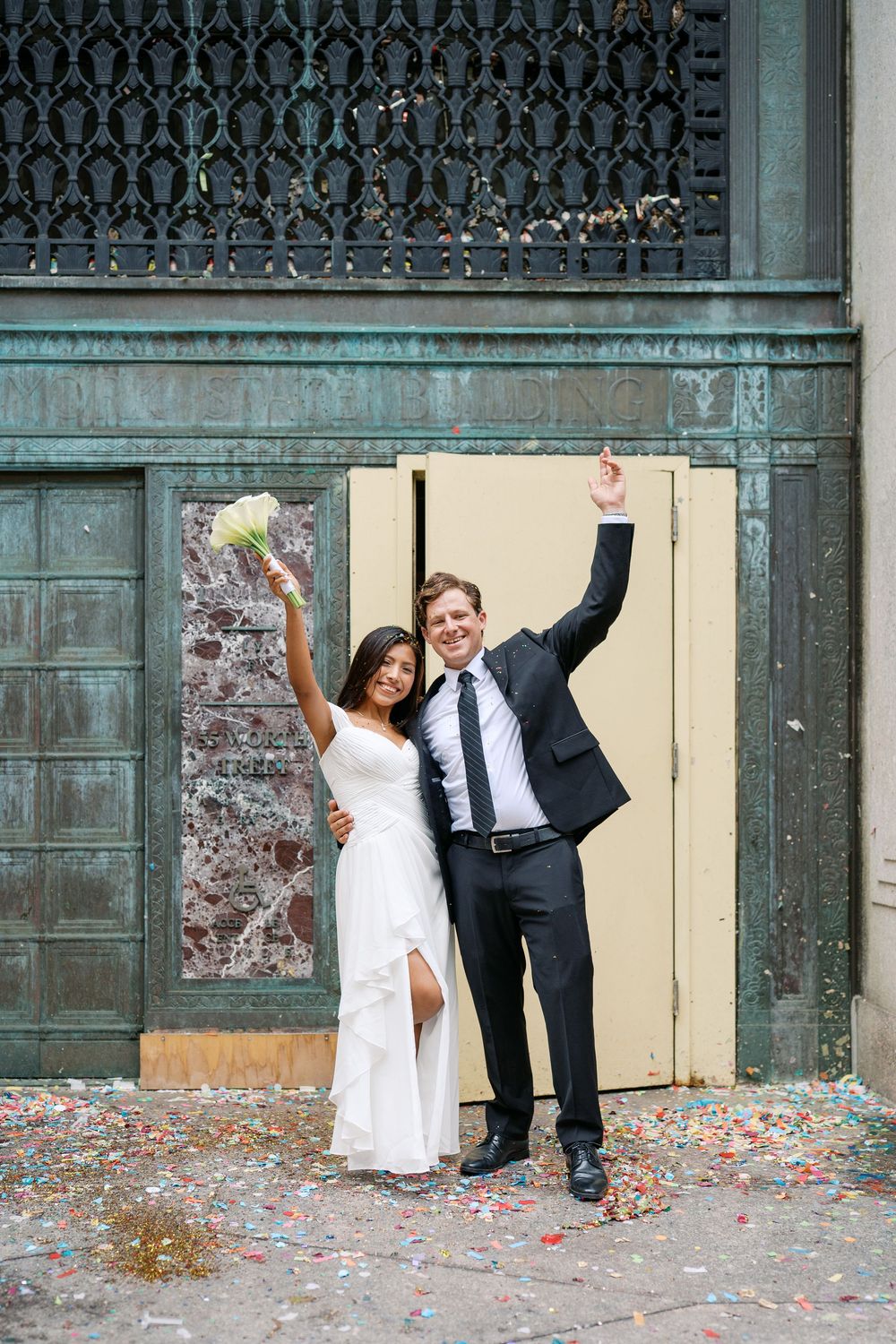 Joyful newlywed couple celebrating with confetti, bride in white dress holding bouquet, groom in black suit raising arms.