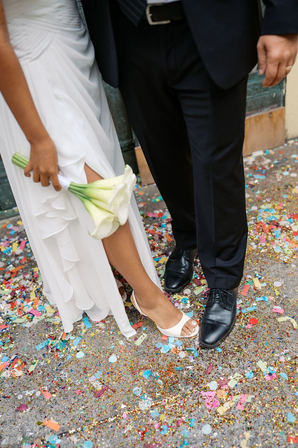 Bride and groom standing on colorful confetti, bride holding white calla lily bouquet wearing white dress and heels.