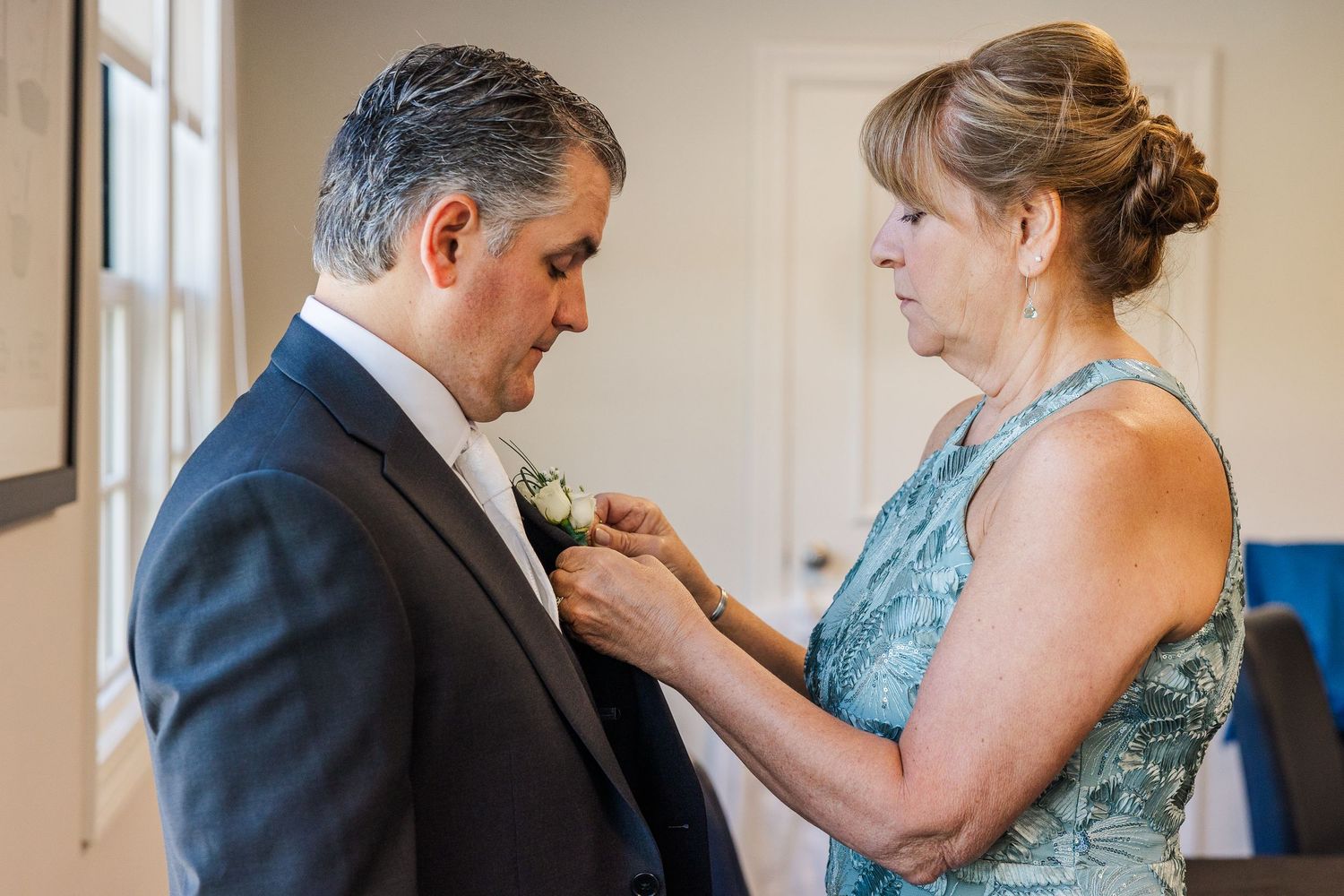 Woman pinning a boutonniere on a man in a suit at a wedding ceremony preparation.