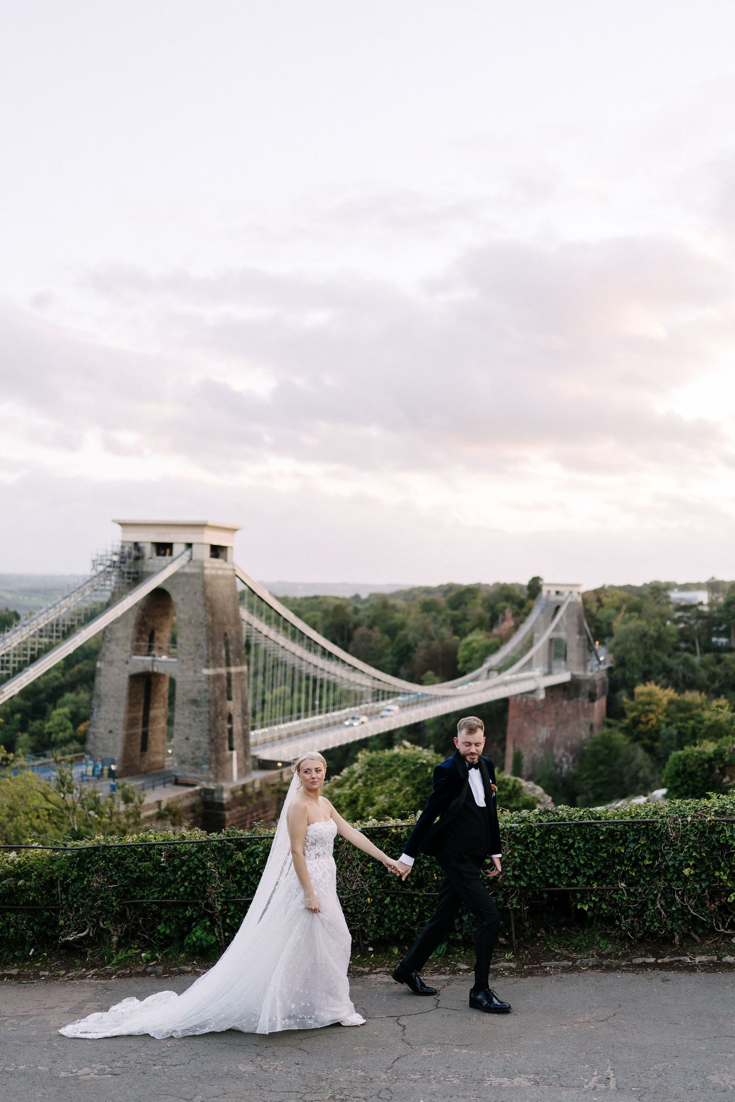 Bride and groom holding hands in front of Clifton Suspension Bridge, Bristol, on their wedding day.