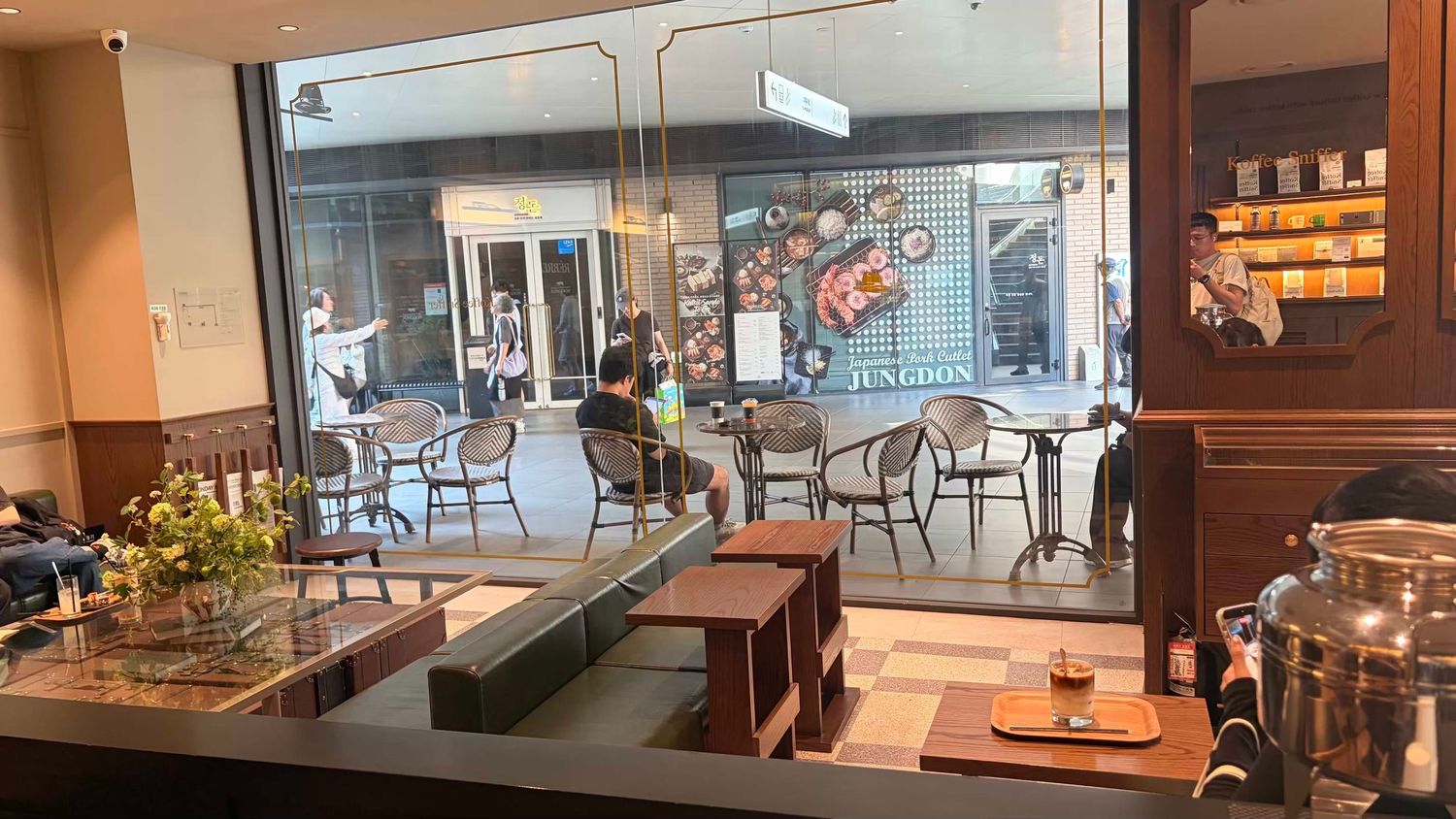 Modern cafe interior with large windows overlooking a bustling street, featuring cozy seating and warm lighting.