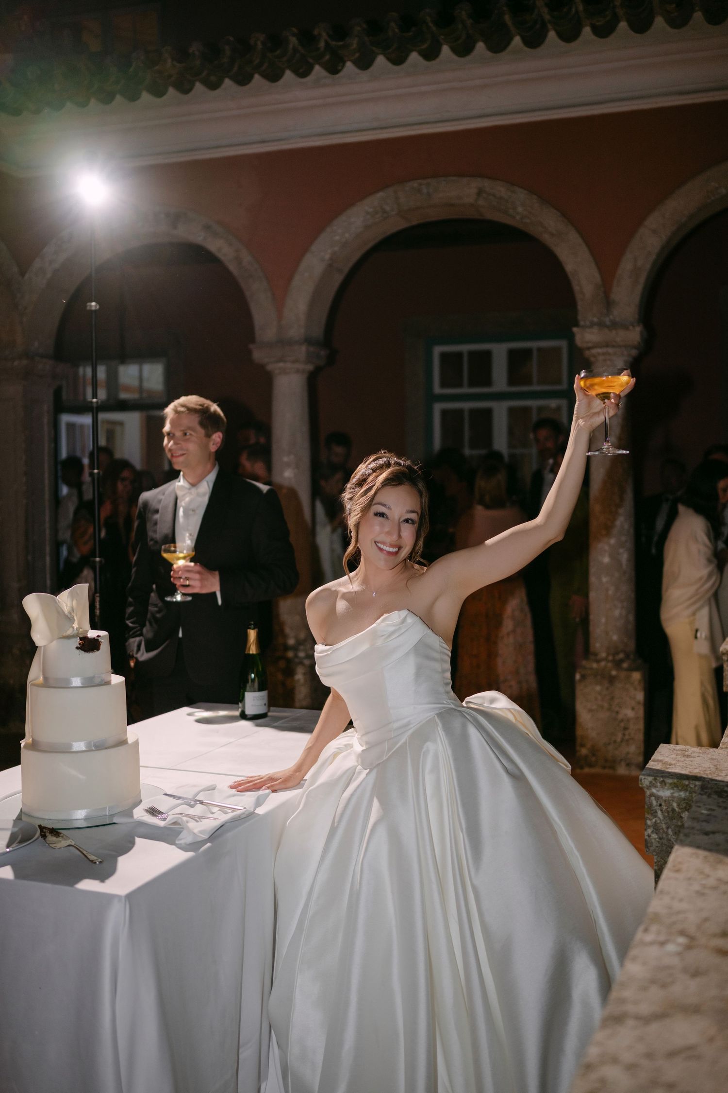 Bride in white strapless gown raises champagne glass beside wedding cake at elegant venue reception.