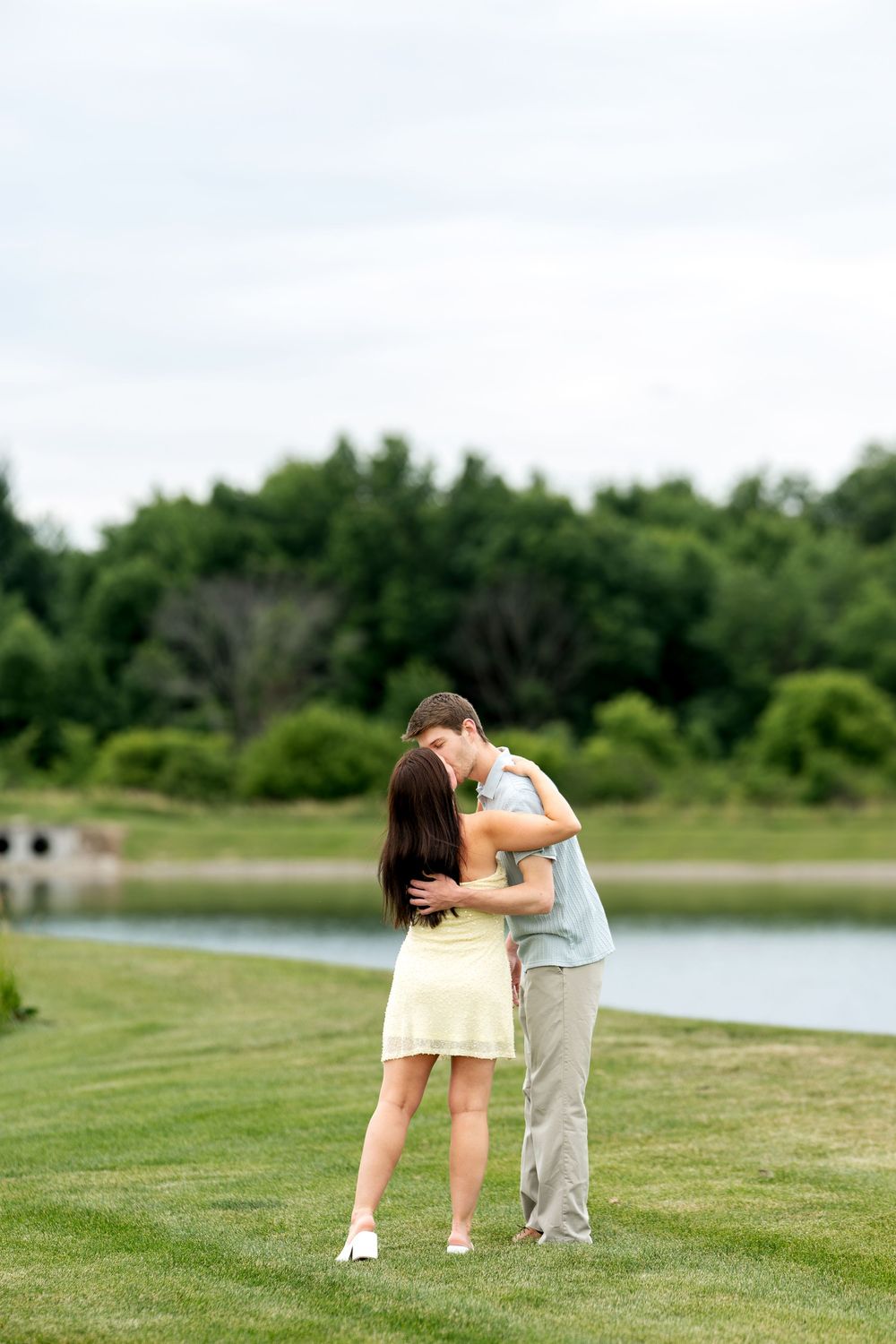 A young couple sharing a romantic kiss on a green lawn beside a peaceful lake surrounded by lush trees.
