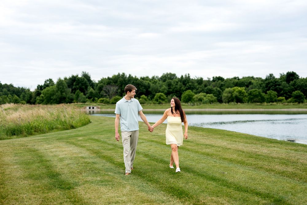 Couple holding hands walking on green grass path beside a calm lake surrounded by lush trees on a cloudy day.