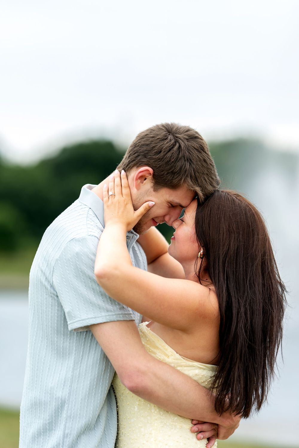 A romantic couple embraces and touches foreheads near a park fountain.