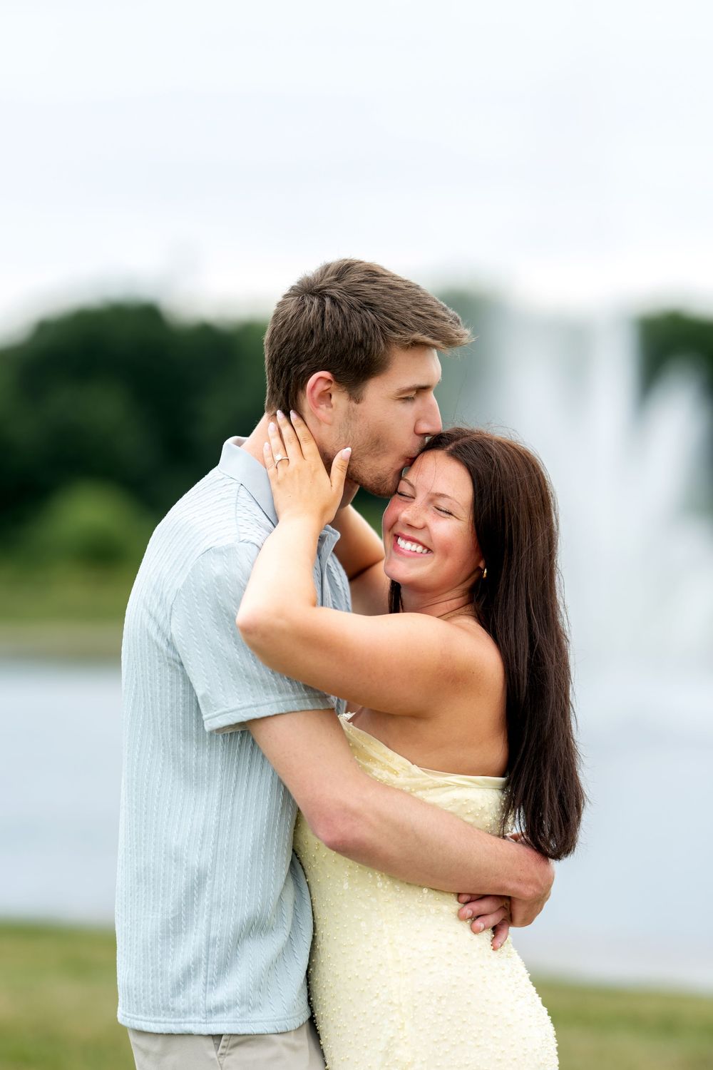 Happy couple hugging and kissing near a fountain in a lush green park setting.