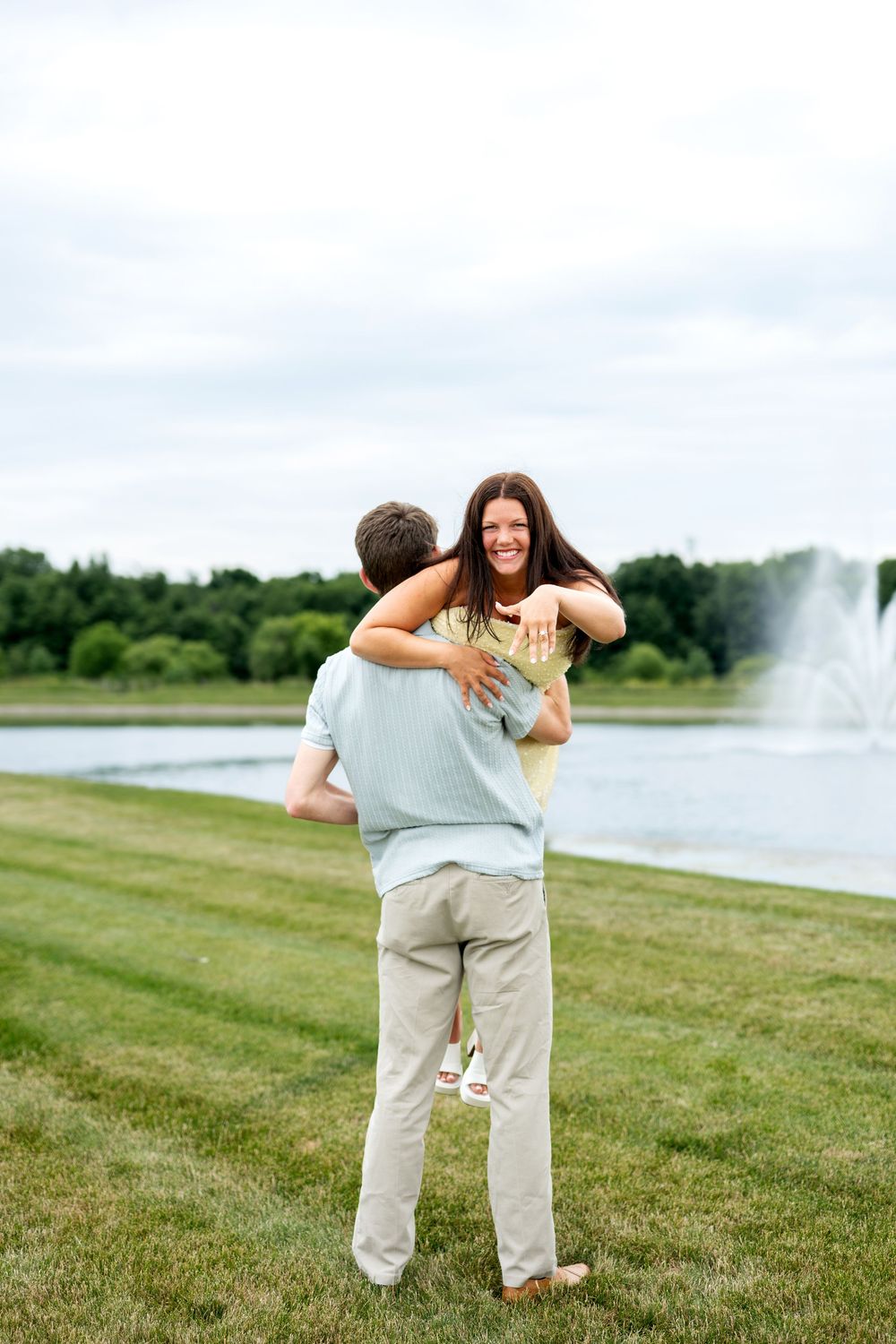 A happy couple embraces and spins together on a green lawn near a pond with a fountain in a park.