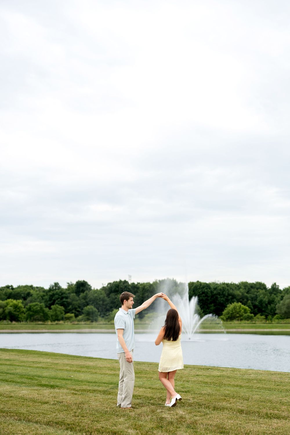 A happy couple dances together on a green lawn by a park fountain.