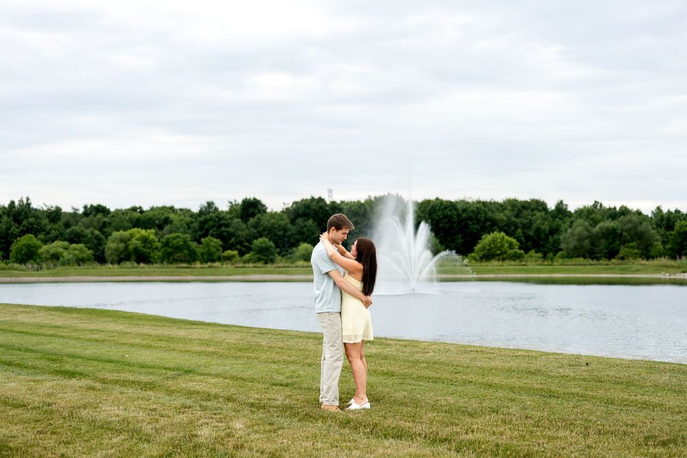 Couple embracing near a serene pond with a fountain and lush green trees in background.