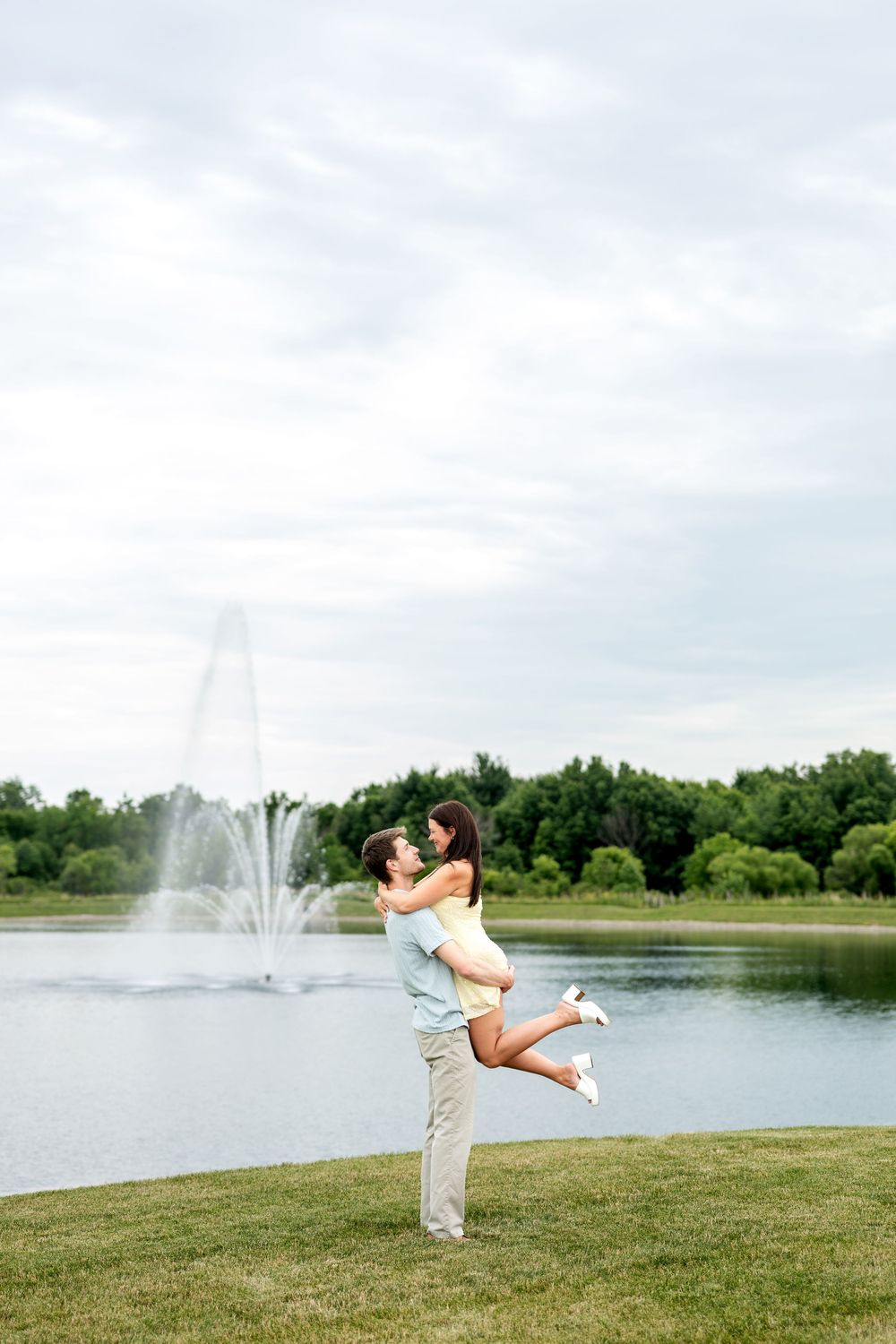 Man lifting woman joyfully in the air near a lake fountain in a green park.