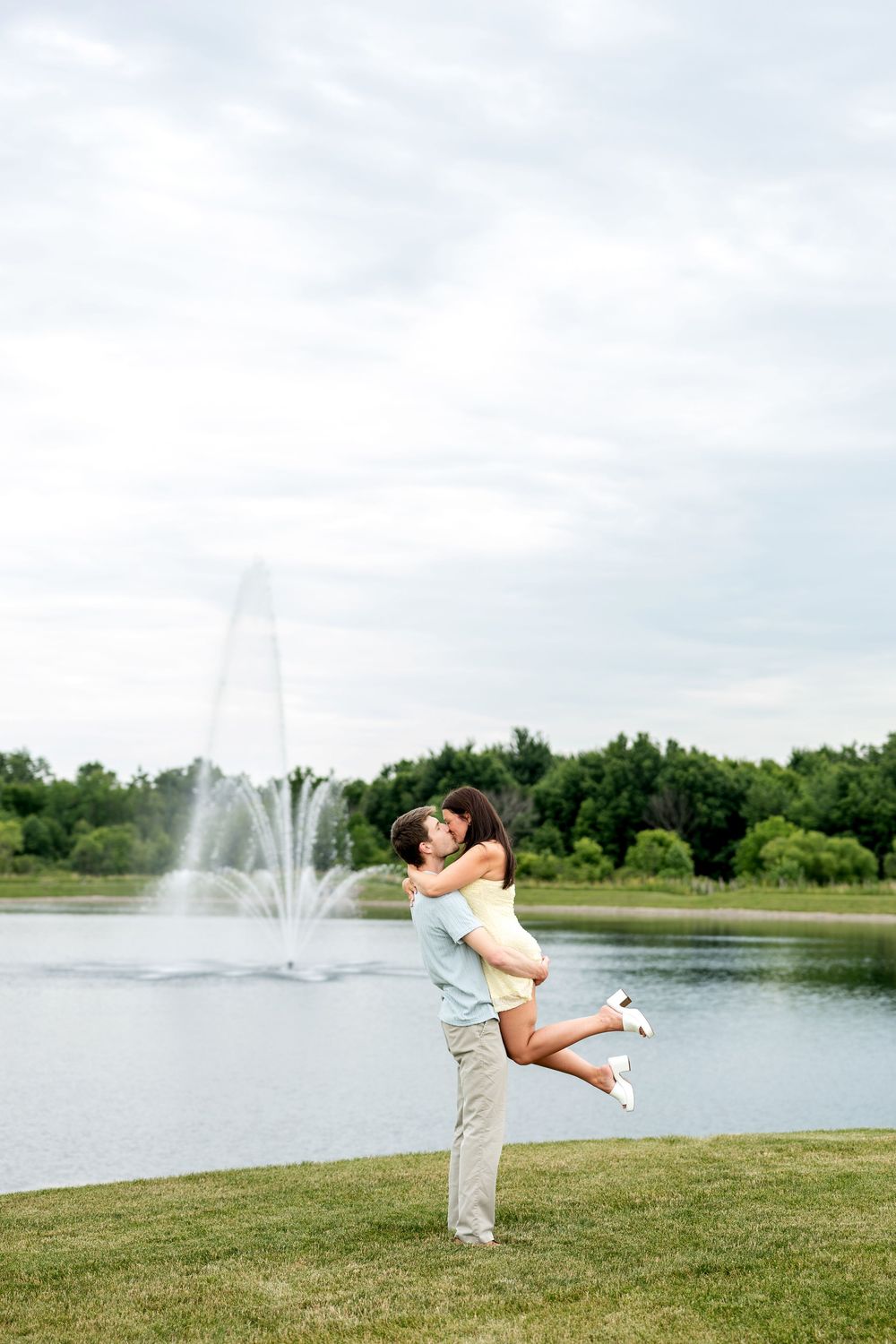 Man lifting woman joyfully by a lake fountain in a green park on a cloudy day.