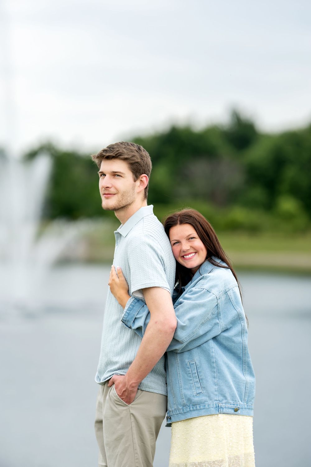 A happy couple poses together outdoors near a fountain, with the woman hugging the man from behind and smiling.
