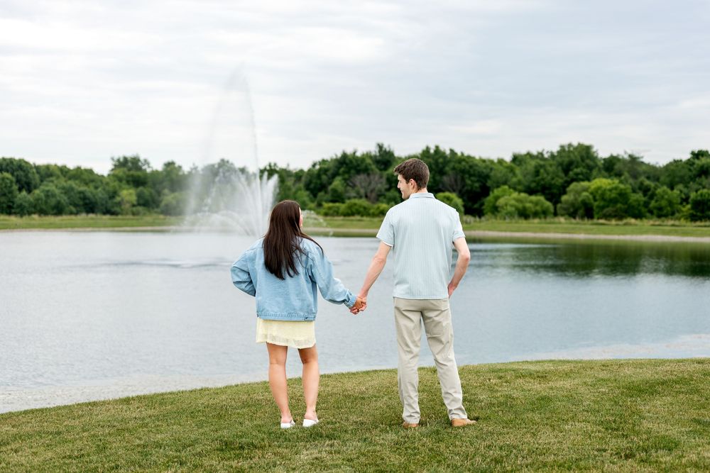 A couple holding hands while gazing at a beautiful fountain on a calm lake in a green park.