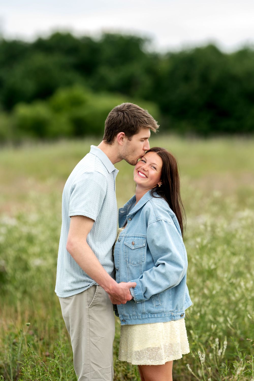 A man kissing a smiling woman on the cheek in a blooming wildflower field on a cloudy day.