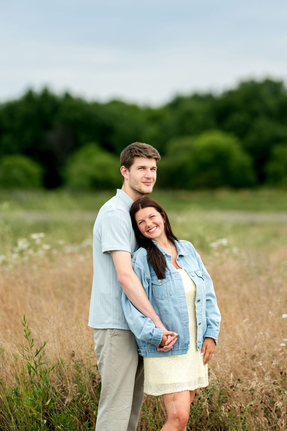 Happy couple embracing in a golden field, woman in denim jacket smiling, green trees behind them.