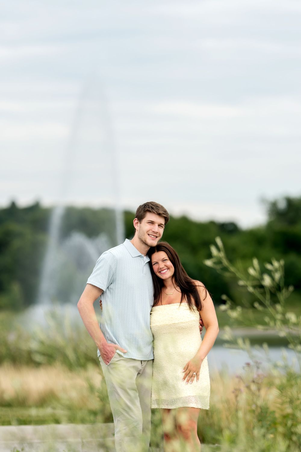 A joyful couple stands together near a park fountain, smiling warmly with lush greenery in the background.