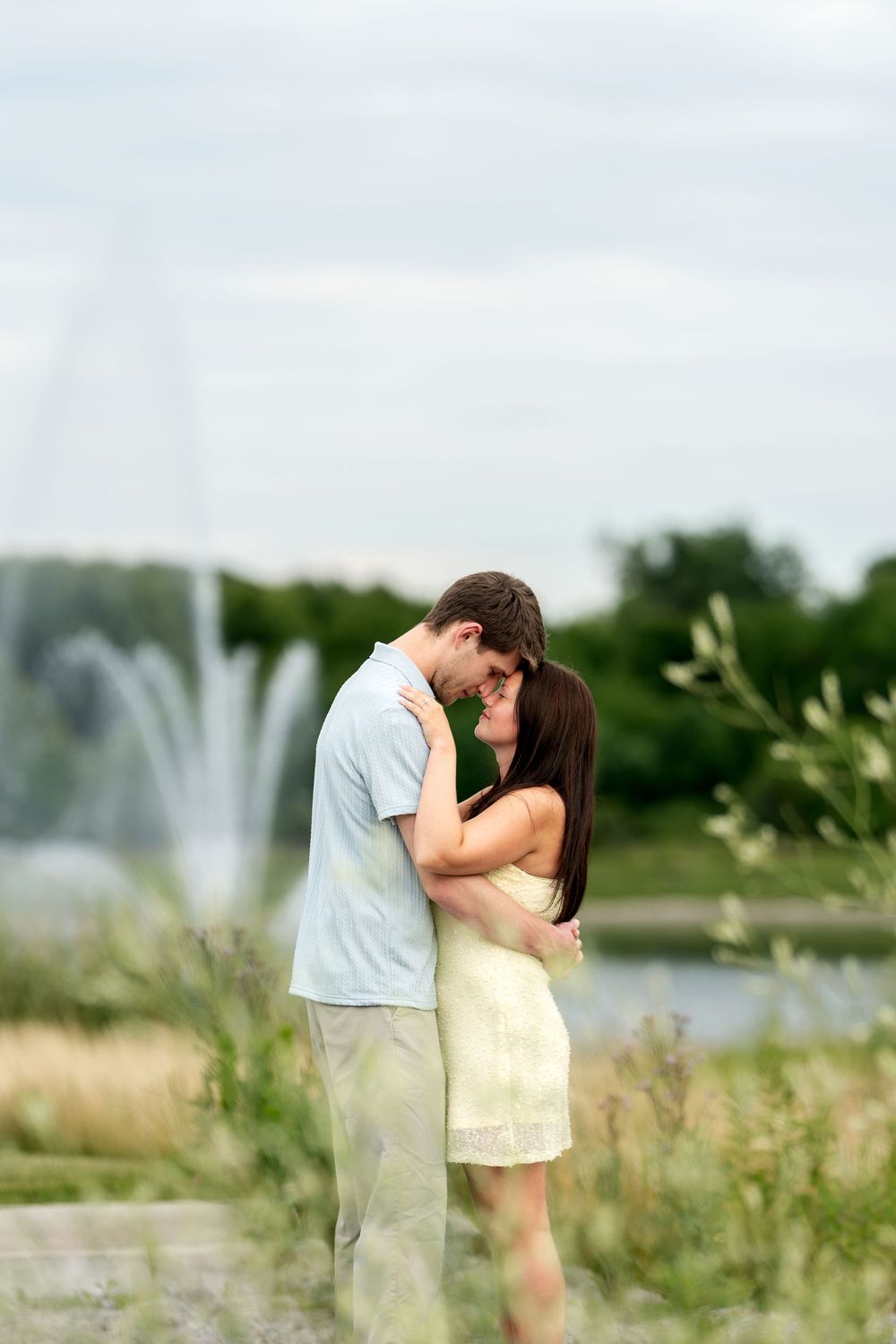 Romantic couple nearly kissing while embracing near a fountain in a green outdoor setting.