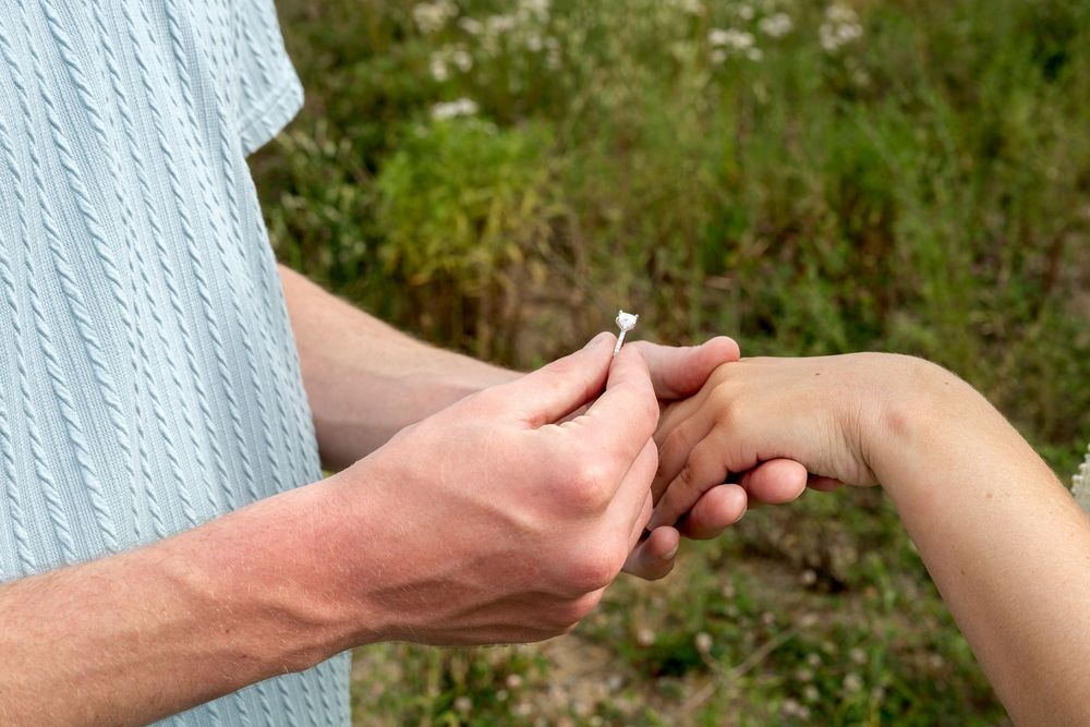A man places a small white flower on a woman's hand outdoors in a green natural setting.