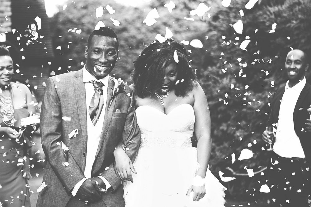 Black and white photo of a joyful couple in wedding attire walking through a shower of confetti. At Oaks farm wedding venue in Surrey.