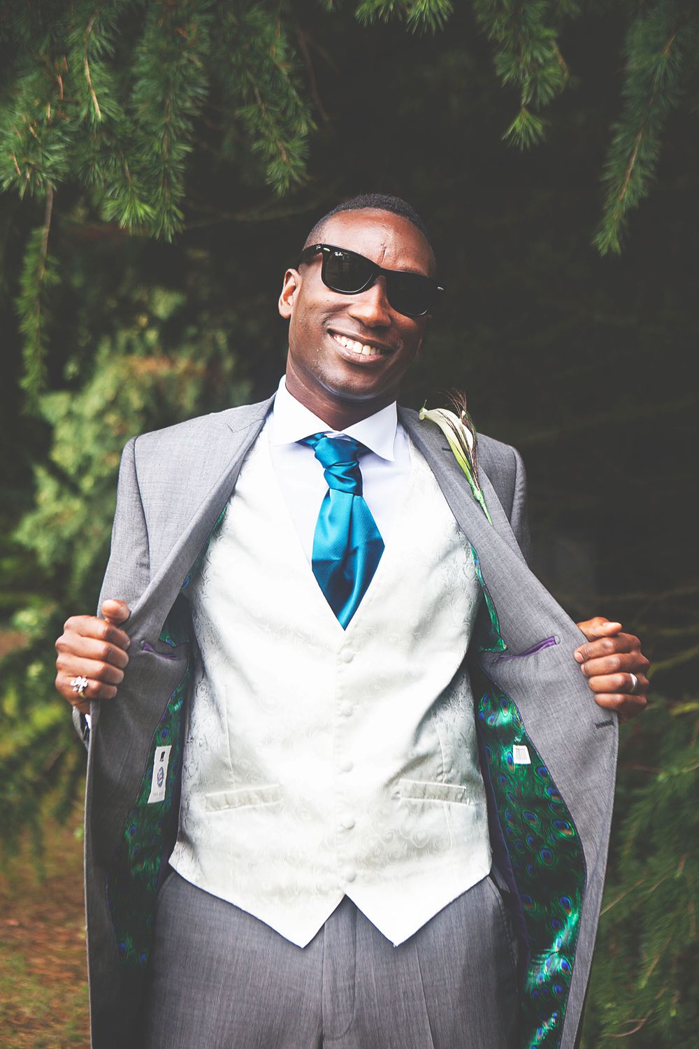 Smiling groom in gray suit, white vest, blue tie, and sunglasses, posing outdoors near evergreen trees. At Oaks farm wedding venue in Surrey