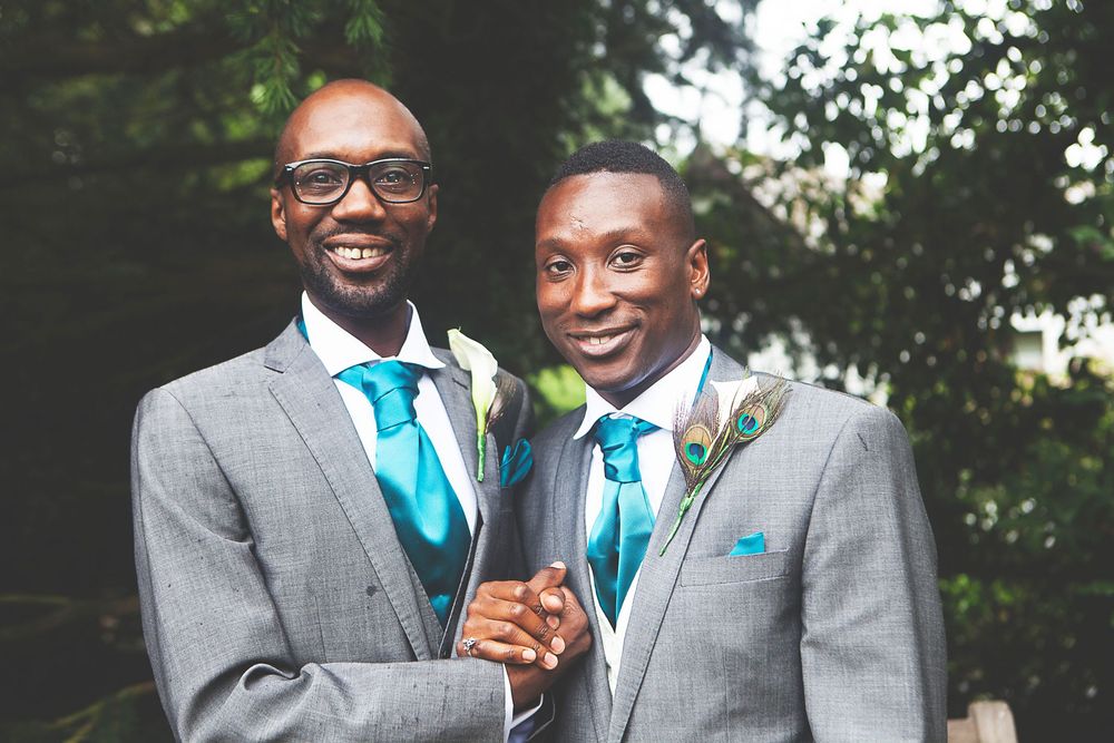 Two men in matching grey suits with teal ties smile and hold hands at a wedding ceremony outdoors. At Oaks farm wedding venue in Surrey.
