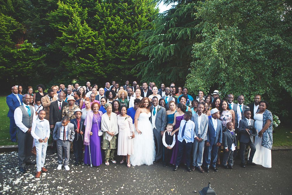 Large wedding group photo outdoors with bride and groom surrounded by guests near lush green trees. At Oaks farm wedding venue in Surrey.