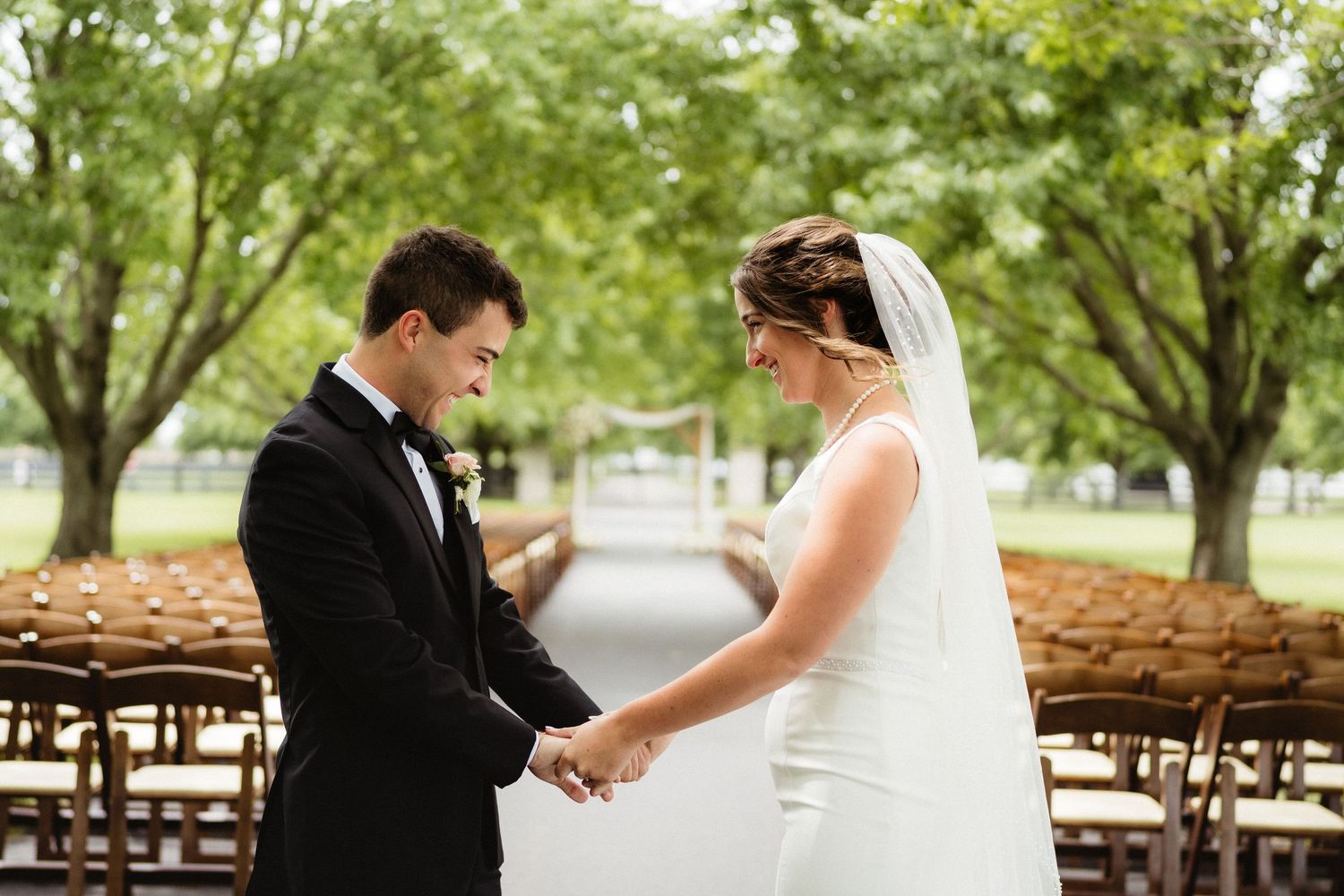 Bride and groom hold hands during outdoor wedding ceremony surrounded by trees and wooden chairs.