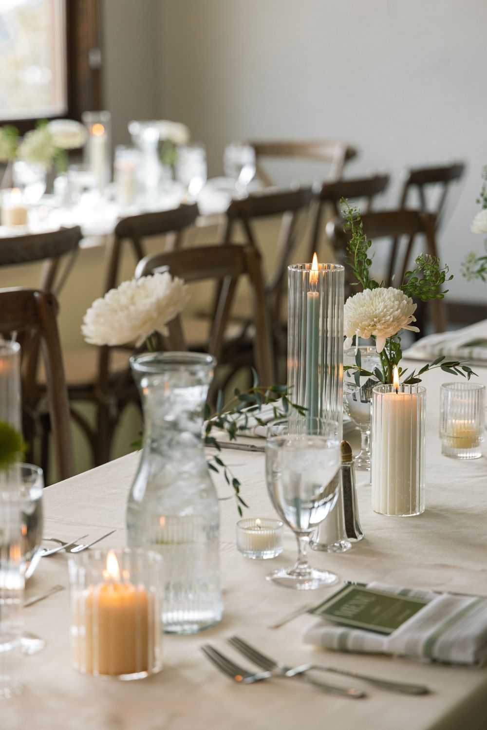 Elegant wedding reception table set with white flowers, lit candles, glassware, and green accents on a marble surface.