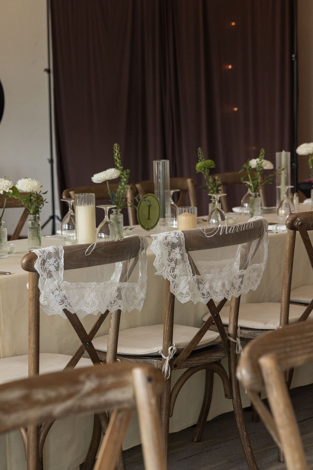 Rustic wedding head table with lace chair sashes, white floral arrangements, candles, and wooden cross-back chairs.