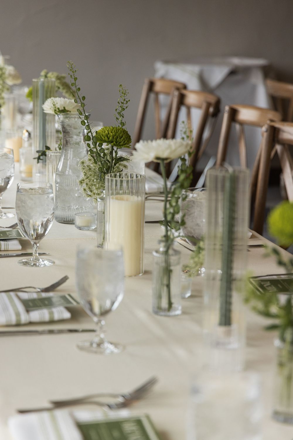 Elegant wedding reception table set with white candles, green floral arrangements, glassware, and striped napkins on a cream tablecloth.
