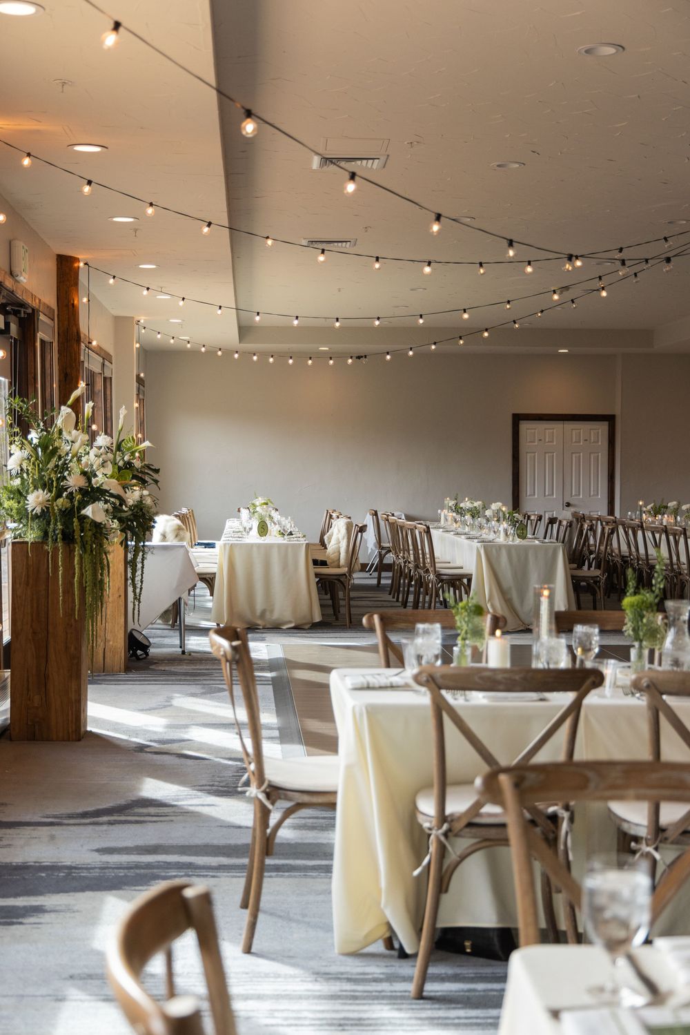 Elegant wedding reception hall with string lights, wooden cross-back chairs, ivory linens, and floral arrangements on round tables.