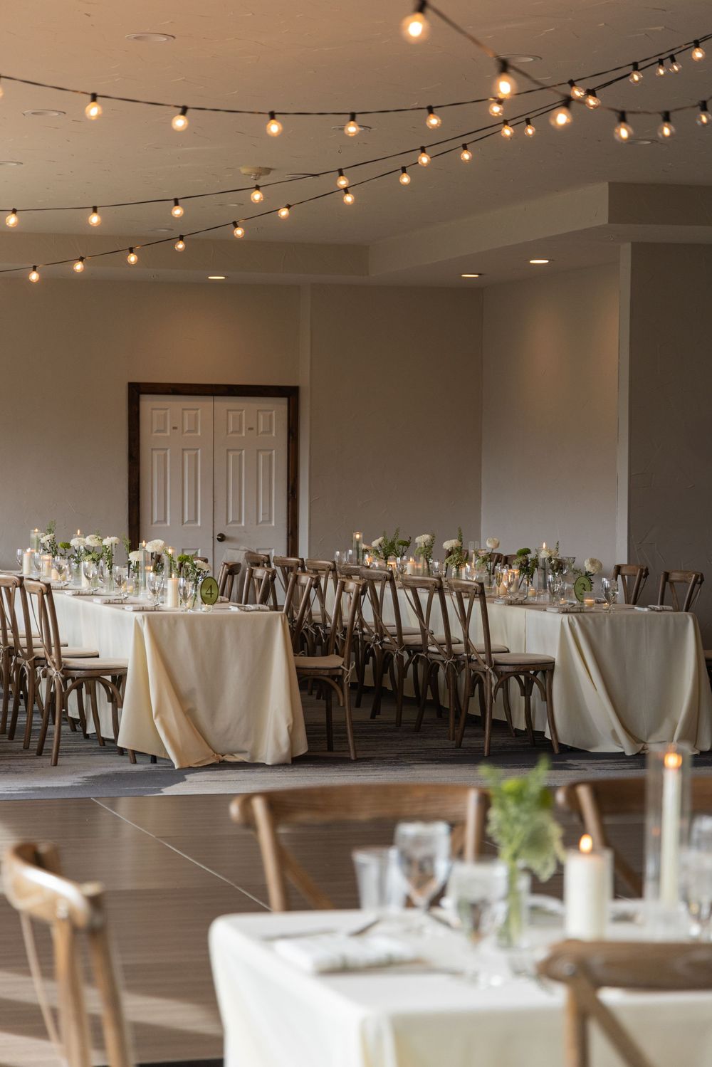 Elegant wedding reception hall with string lights, long tables draped in ivory linens, wooden cross-back chairs, and candle centerpieces.