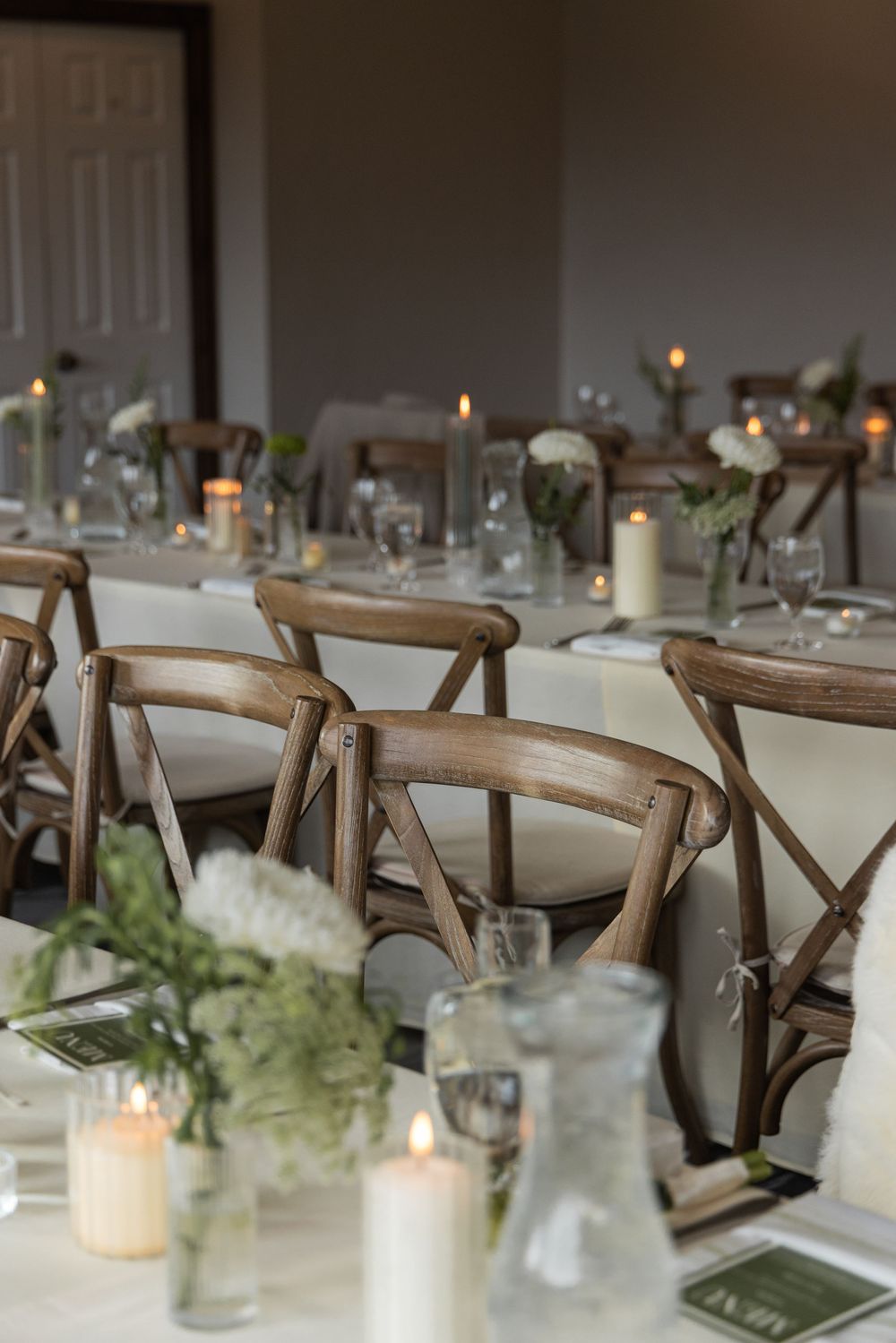 Elegant wedding reception tables with wooden cross-back chairs, white candles, floral centerpieces, and soft candlelight.