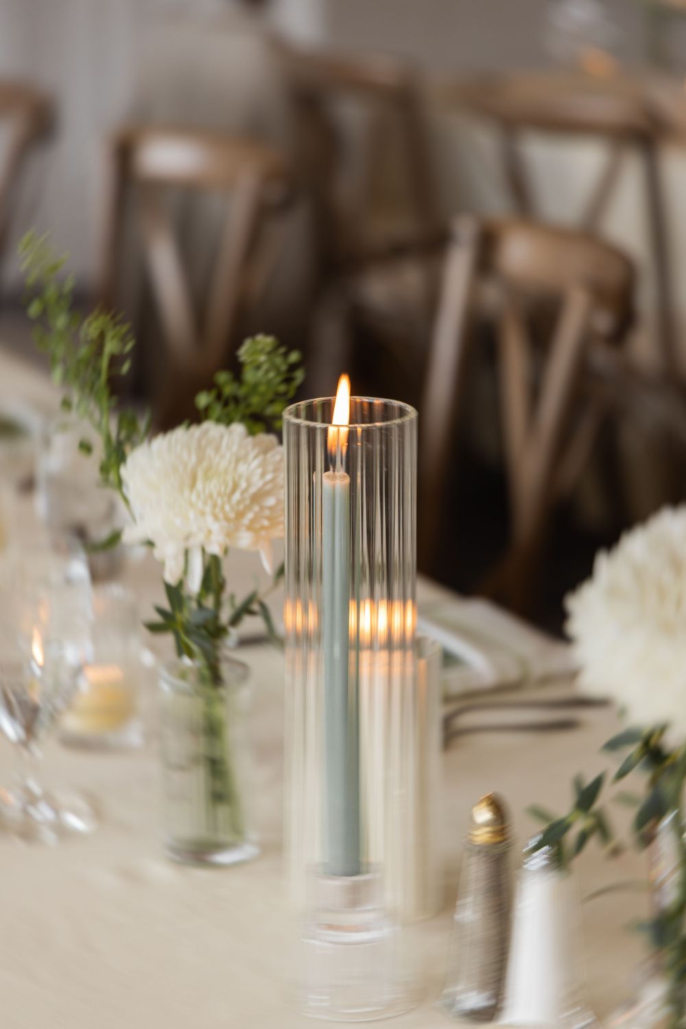 Elegant wedding table setting with lit pillar candle in glass holder, white flowers, and wooden cross-back chairs.