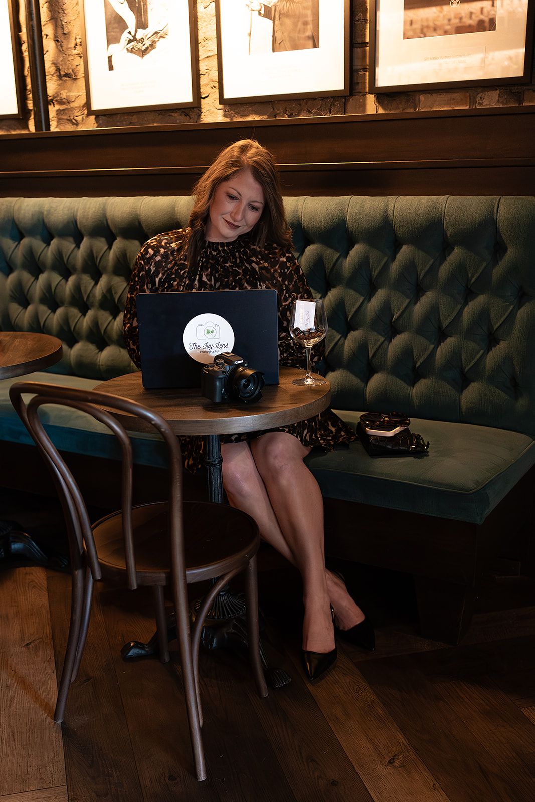 Woman in leopard print dress working on laptop at elegant restaurant booth with wine.
