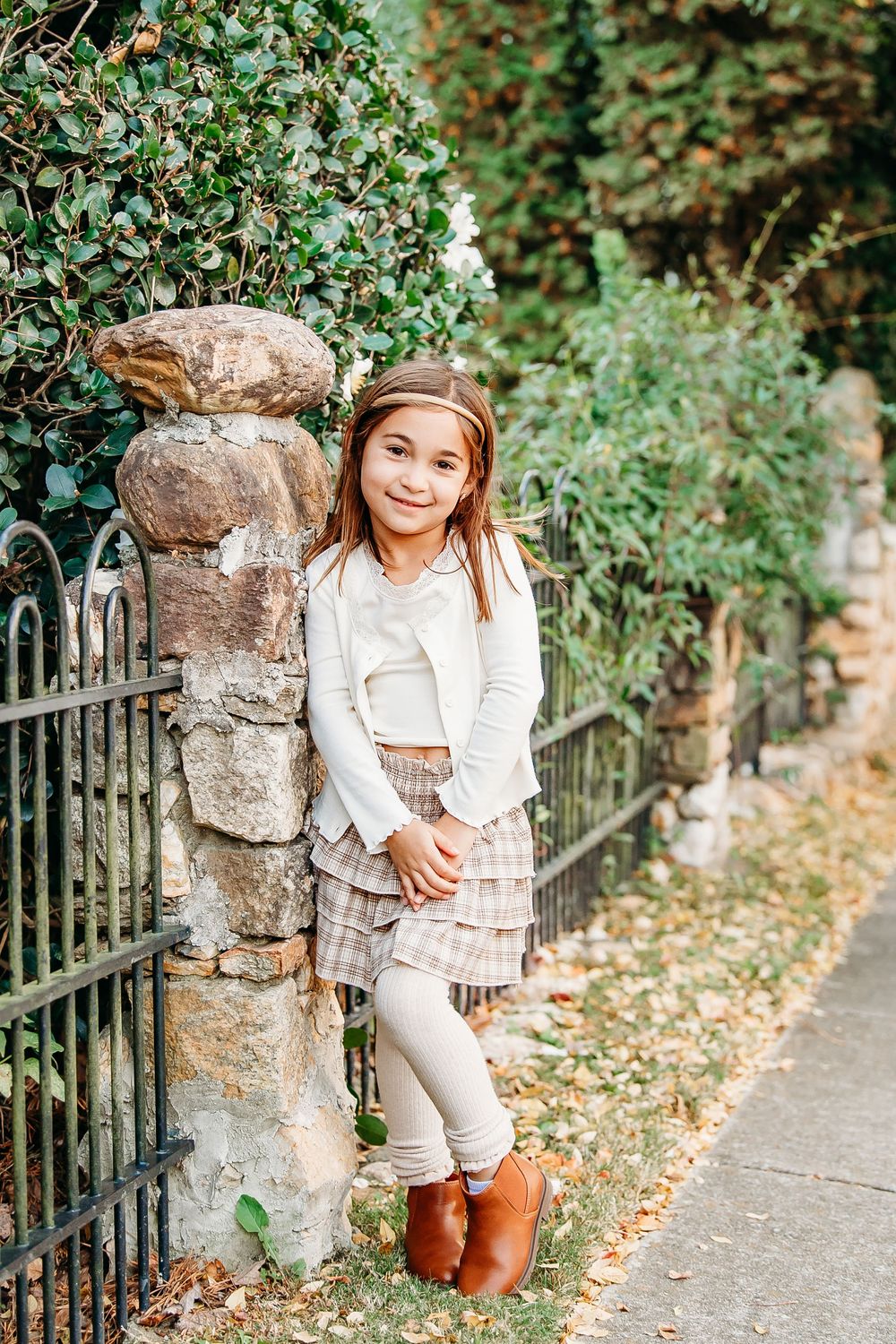 A young girl in a plaid skirt and white top smiles while leaning against a stone fence outdoors.