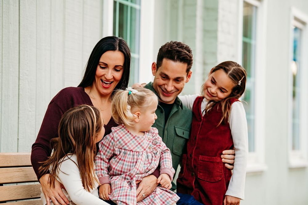 A joyful family of five laughs together while sitting on a bench outside a white building.